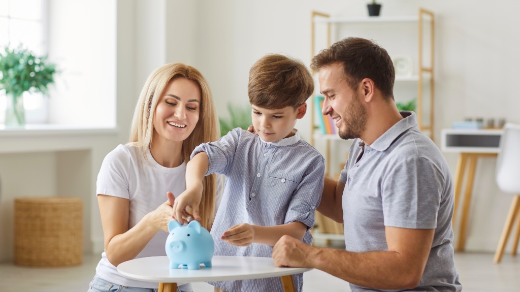 Parents and child together put a coin into a piggy bank, teaching savings and financial habits. This family moment of saving money occurs in home, showcasing unity and planning together.
