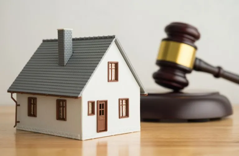 Small white model house with gray roof and brown window frames next to a wooden judge's gavel.