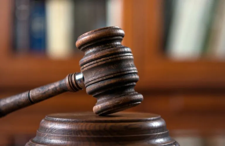 Close-up of a wooden judge's gavel resting on a sound block with blurred bookshelves in background.