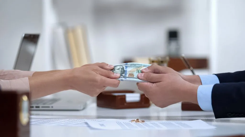 Two people exchanging a stack of US hundred-dollar bills over a desk with documents and wedding rings.