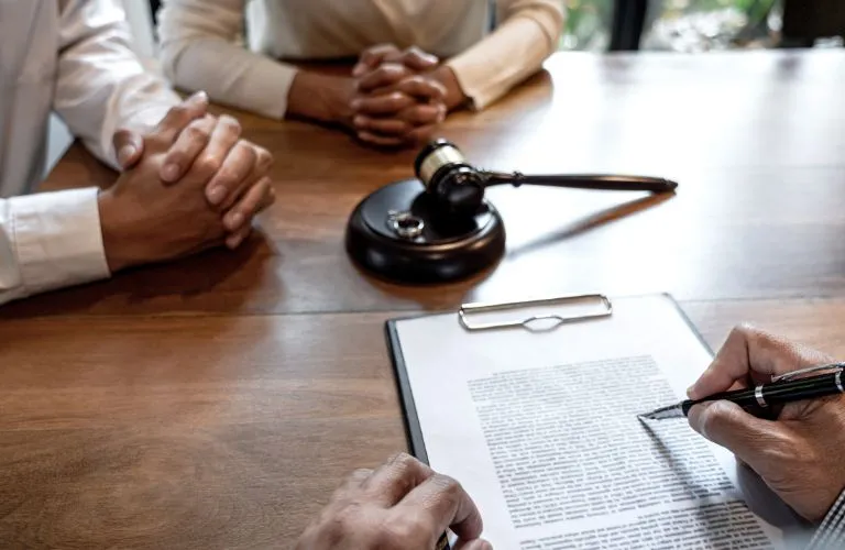 Two people with folded hands opposite a person signing a document, with a judge's gavel on the table.
