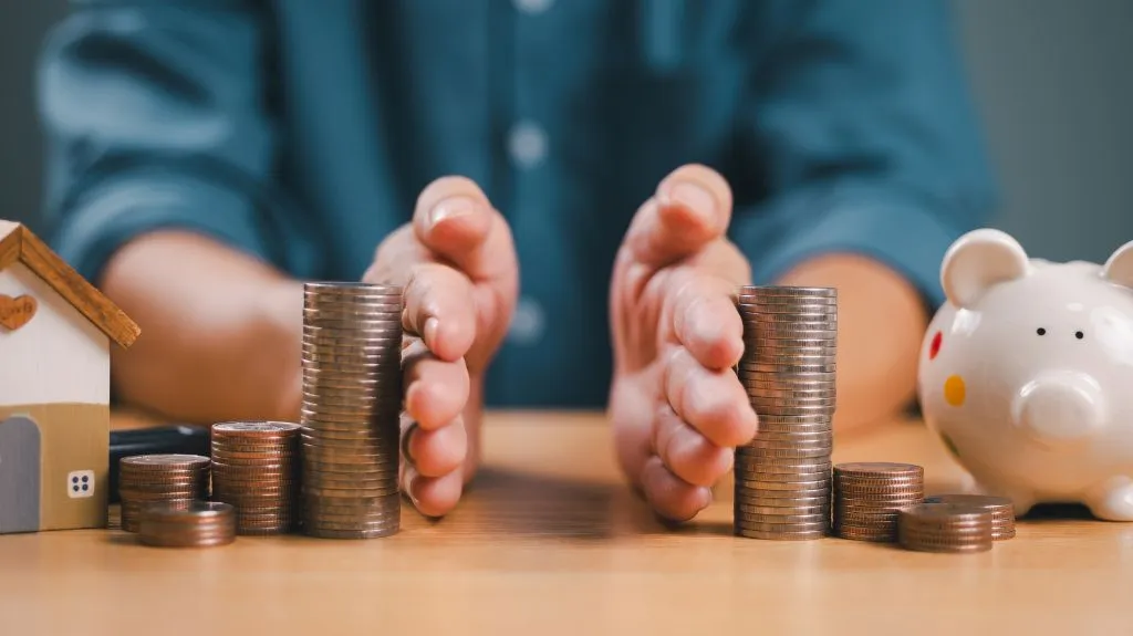 Hands separating stacks of coins on a wooden table between a small house model and a white piggy bank.