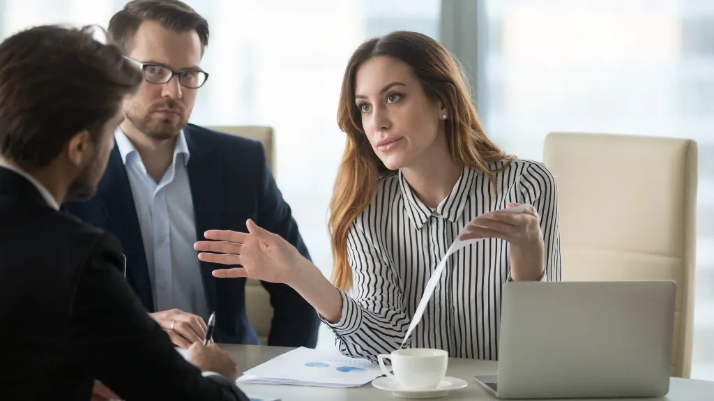 Three business professionals engaged in a discussion, woman gesturing with document, laptop and coffee cup on table.
