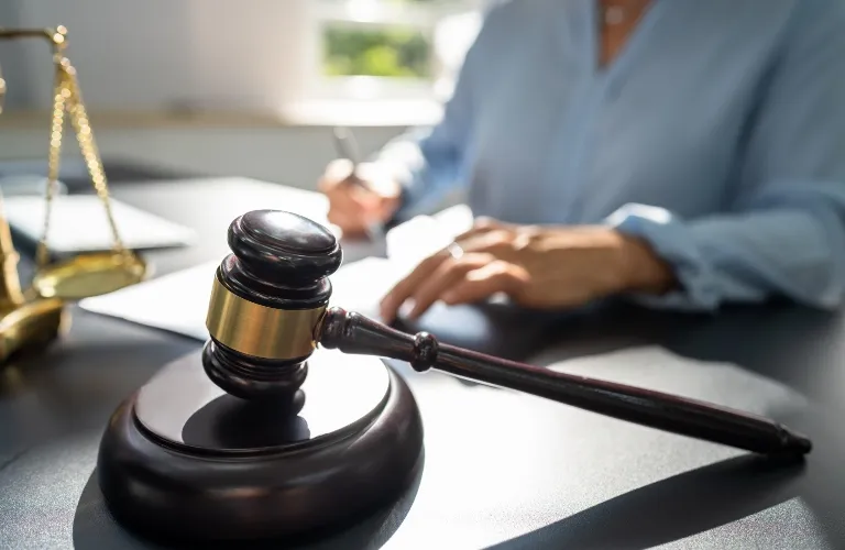 Dark wooden judge's gavel with brass band on desk, blurred person writing in background.